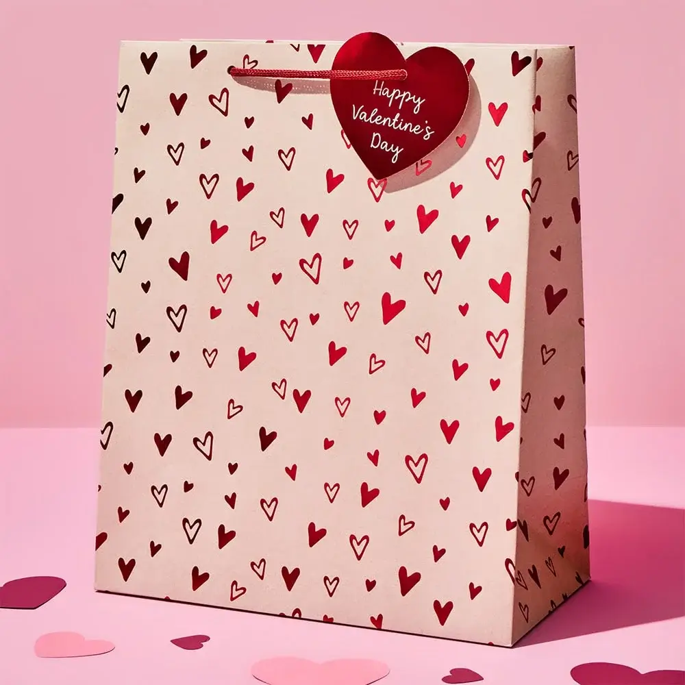 Valentine’s Day gift bag with red and pink heart patterns and a red heart-shaped tag that says Happy Valentine’s Day, placed on a pink surface with scattered paper hearts.